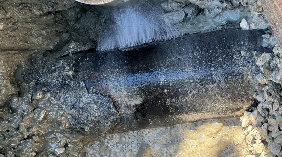 Close-up of an active underground water main leak, with pressurized water spraying from a damaged pipe section during excavation and leak detection investigation.